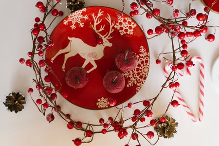 A red plate with a Christmas pattern is on the table, on it are red small apples, next to a branch with red berries. Christmas flat layの写真素材