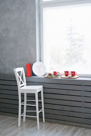 A white wooden bar stool stands near the windowsill and window, on which are Christmas red mugs and decorの写真素材