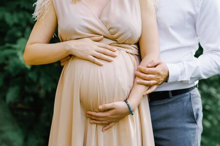 A pregnant woman in a beige long dress holds her hands on her stomach as her husband and baby daddy-to-be hugs his wife. Close-up of arms and abdomen. Pregnancy womenの写真素材