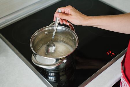 A woman's hand stirs the sugar syrup liquid in a saucepan on an electric stove with a tablespoonの写真素材