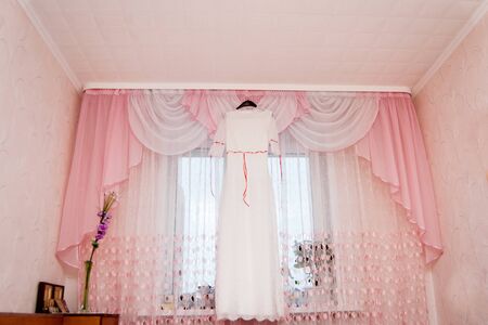 A white wedding dress hangs on a window in the living room. Clothing for the brideの写真素材