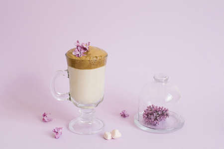trendy drink 2020 dalgon Coffee in a transparent Cup with a paper tube, a plate with lilac flowers on a lilac background with a copy spaceの写真素材