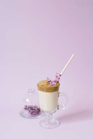 dalgona Coffee in a transparent Cup with a paper tube, a plate with lilac flowers on a lilac background with a copy spaceの写真素材