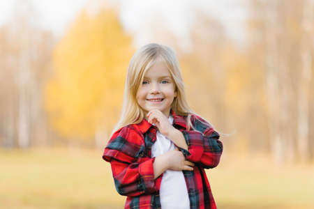 Close-up portrait of a cute charming smiling little blonde girl standing in an autumn autumn Park outside looking at the camera, happy lifestyle concept of childhoodの写真素材