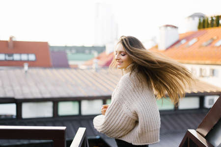 A beautiful young brunette is smiling broadly, her hair developing in the wind as she spins around. Portrait of a pretty female model in a fashionable knitted beige sweater posing while walking along a city street.の写真素材