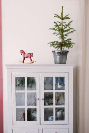 Christmas tree in a metal bucket, vintage wooden red toy horse on a white dresser in the interior of...