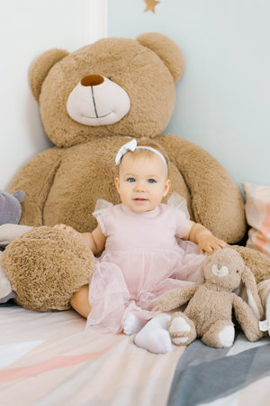 A cute one year old girl is sitting on the bed in the children's room at home among soft funny toys in a pink dressの写真素材