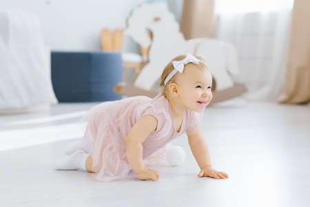 A pretty blue eyed blonde one year old in a pink dress crawls on all fours in the nursery on the...