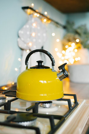 A yellow teapot stands on the stove in the kitchen against the background of Christmas lightsの写真素材