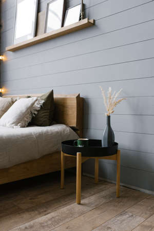A black bedside table with a gray vase and dried flowers and a green metal mug placed by the bed in...