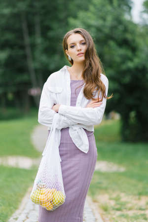 A young woman in white lilac clothes with an eco-bag with fruit on the background of nature. Plastic-free conceptの写真素材