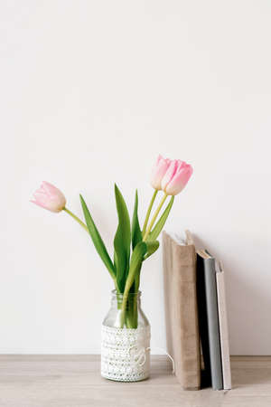 A bouquet of three pink tulips stands in a glass vase with lace. Next to it are notebooks or notepads and a mug of tea, a photo album in a velor fabric cover of beige colorの写真素材