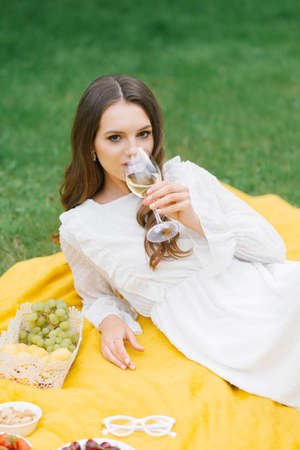 A beautiful young woman lies on a yellow blanket on the grass, holding a glass of wineの写真素材