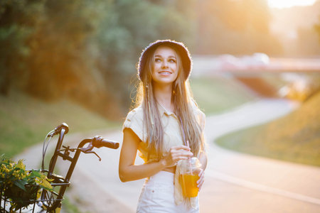A happy girl in white shorts and a yellow blouse holds a glass of juice with a straw on the background of the road near the forestの写真素材