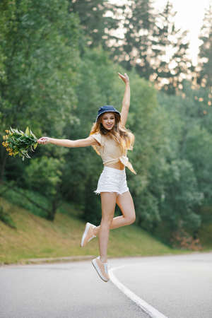 A happy girl in white shorts and a yellow blouse jumps up with a bouquet of flowers, standing on the road near the forestの写真素材