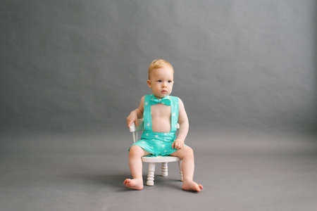 A funny little boy in a summer suit is sitting on a white children's chair on a gray background in a photo studioの写真素材