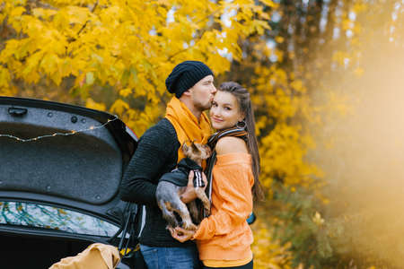A guy and a girl in love embrace in an autumn Park, a guy holds a cute dog in his arms and kisses his favorite girlの写真素材