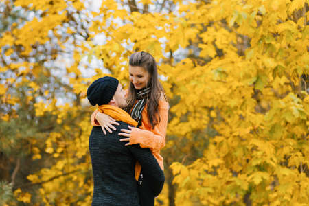 A young couple in an autumn Park. A smiling man holds a woman in his arms against a colorful autumn background. Happy couple on the background of autumn trees. A couple having fun in the autumn Park.の写真素材