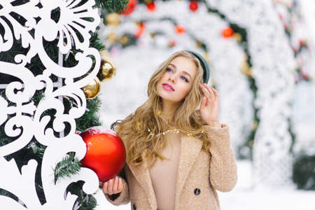 Street photo of a young beautiful happy smiling girl with a red Christmas ball in her hands, posing on the street. A festive Christmas market in the background. The model is dressed in a stylish winter coatの写真素材