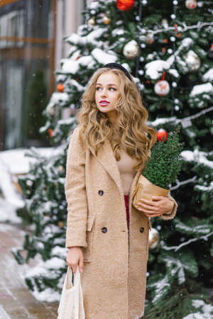 A pretty blonde holds a potted Christmas tree in her hands, which she bought in a store, and walks...