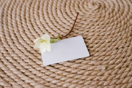 A white blank mock-up of a guest card on a jute rope background and a white delicate flower next to...