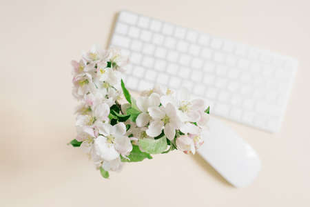 A white vase with a beautiful bouquet of branches of a blossoming apple tree on a beige table and a keyboard with a mouse in decoloration, top view.の写真素材