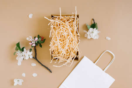 Mockup of an open gift box with peach colored paper shavings, white paper bag and apple flowers on a beige background. Flat lat, top viwの写真素材