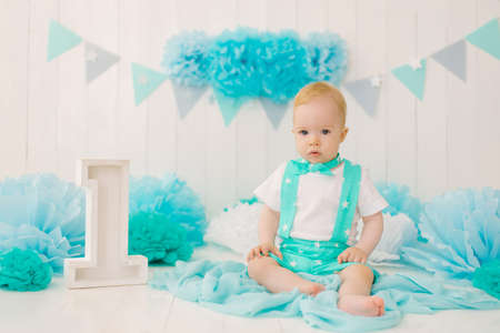 A beautiful one year old boy in turquoise clothes is sitting near the number 1 in the photo zone for...