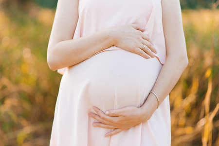 A pregnant woman in a pink delicate dress holds her hands on her stomach, hugs him
