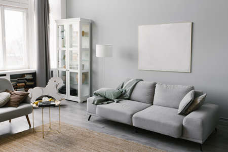 Stylish minimalistic interior of the living room in grey. Sofa with plaid, floor lamp, beige carpet and mockup white frame on the wallの写真素材