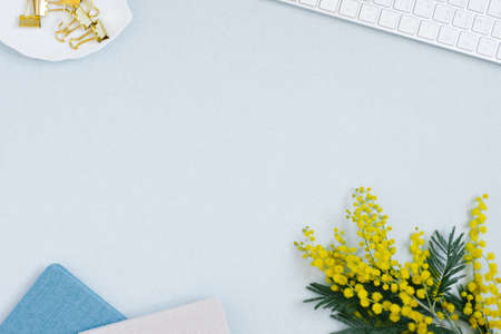On a blue flat lay background with a yellow mimosa flower, keyboard notepad and stationery clips, a...