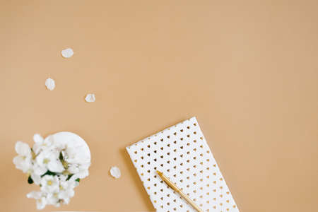 A white notebook with a gold pen on a beige table and flowers in a defocus vase. Blogger's workplace. top view, copy spaceの写真素材