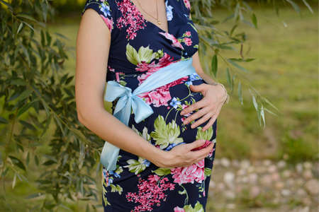 A pregnant woman in a bright colored dress holds her hands on her stomach while waiting for a baby