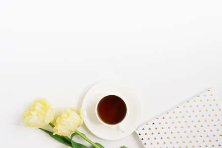 A white porcelain cup with black tea on a saucer surrounded by spring yellow tulips, a notebook on a...