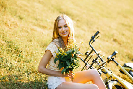 portrait of a beautiful girl in a field, sitting on the grass, next to a bicycle, with a bouquet of...