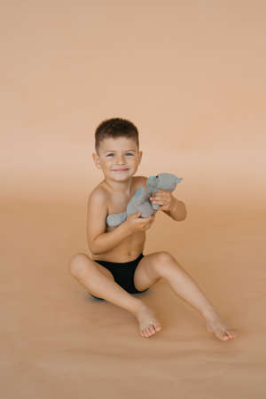 A child boy in underpants holds a soft bear toy in his hands, sitting on a beige background in the studio and smilingの写真素材