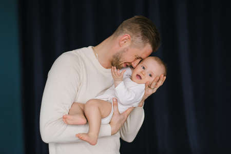 The young man is happy and holds his six-month-old son in his arms and kisses him and hugs him in...