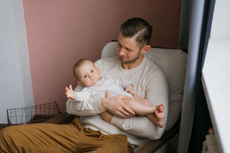 A young Caucasian father holds his sweet sleep baby in his arms, sitting in a chair at homeの写真素材