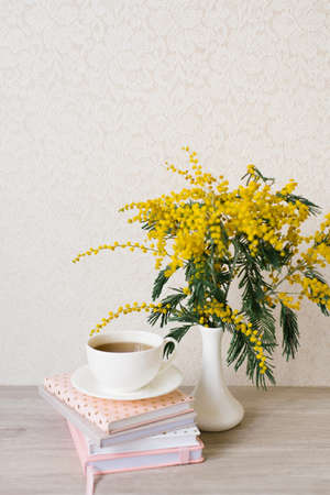 A beautiful composition of mimosa branches and a white tea cup with a saucer and a stack of notebooks on a table with a copy spaceの写真素材