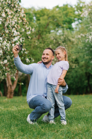 Dad and his six-year-old daughter take selfies on the phone, smiling and having fun on camera in the park in summer or springの写真素材