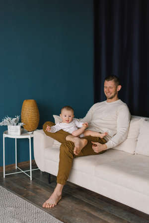 The young man is happy and holds his six-month-old son in his arms, sitting on the sofa in the room