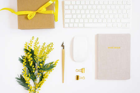 A women's workplace with a computer keyboard, mouse, notepad, pen for calligraphy, paper clips, a gift box with a bow, mimosa flowers on a white background. Geometric flat layの写真素材