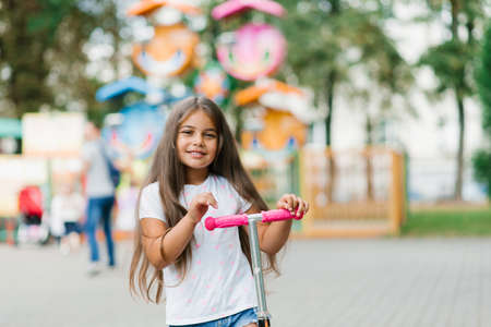 Beautiful smiling girl rides a scooter along the path of an amusement park on a summer day. Seasonal children's active sport.の写真素材