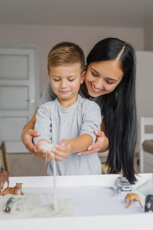 Happy mom and her son play with kinetic sand, developing fine motor skills at home. A smiling family...