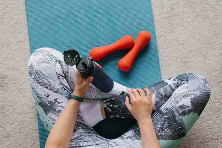 Over the shoulder, a close-up view of a sporty woman sitting on a rug, holding a bottle of water in...