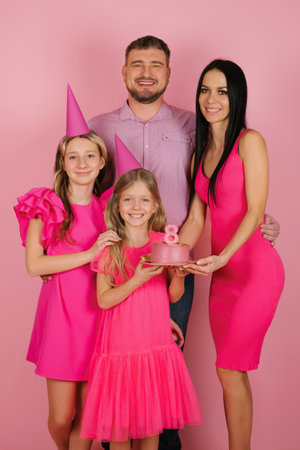 Portrait of a beautiful happy family with an 8-year-old birthday girl with a cake. Mom, dad two sisters in pink dresses with festive hats on their headsの写真素材