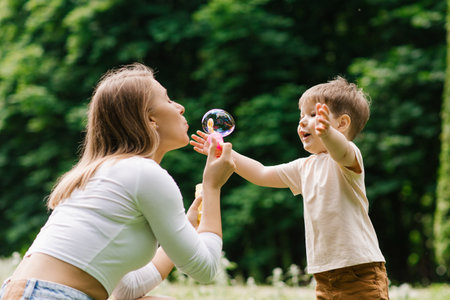 Loving mother plays with her three-year-old son on the lawn and teaches him to blow soap bubbles in a summer parkの写真素材