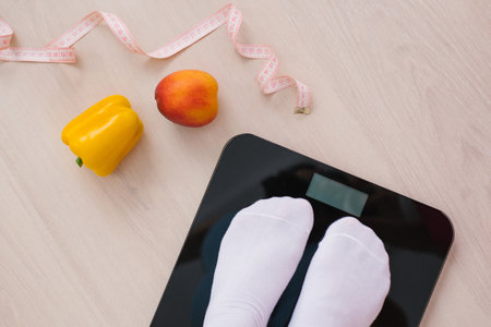 Pair of female feet in white socks standing on a scale with vegetables and fruits and a measuring tape or tape near themの写真素材