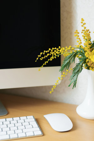 Bouquet of yellow mimosa flowers in a vase on a desktop with a computerの写真素材