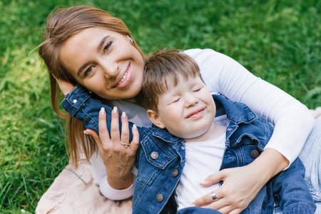 Young Caucasian mother and her young son lie on a blanket on the grass in the park in summer and have fun outdoorsの写真素材